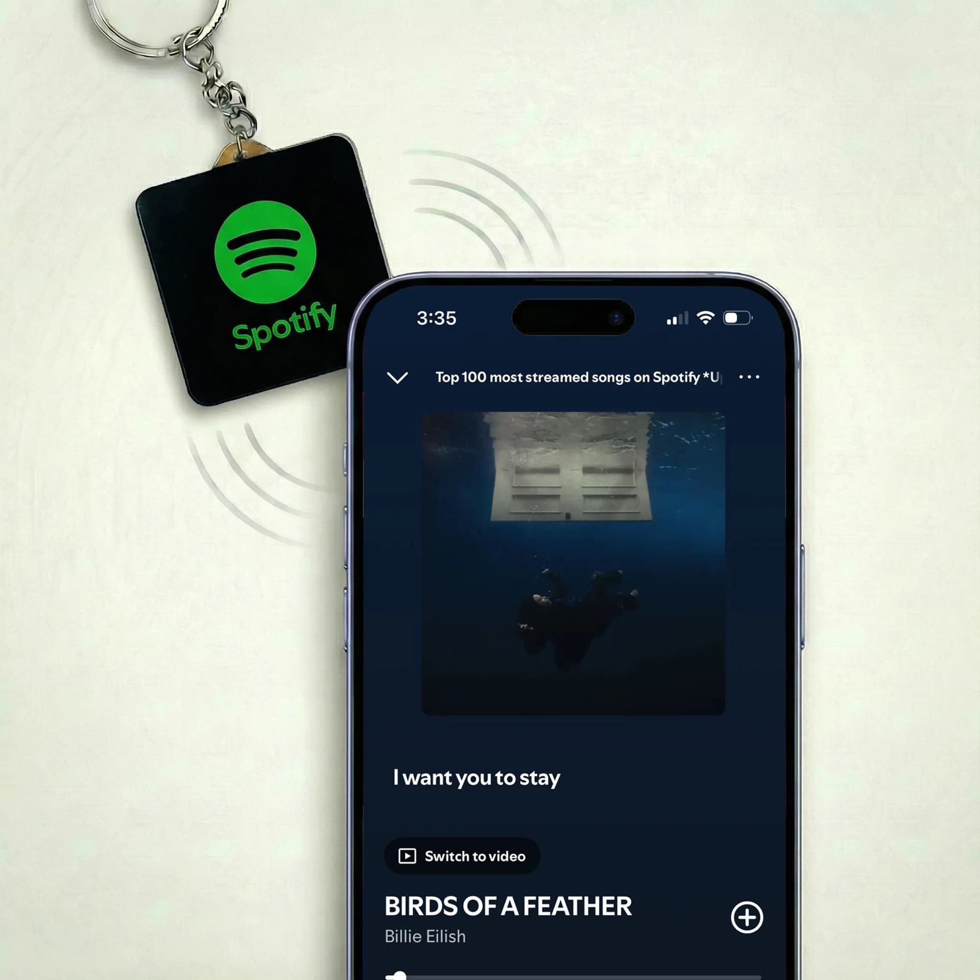 A black square keychain with a green Spotify logo hangs next to a smartphone displaying a Spotify playlist.