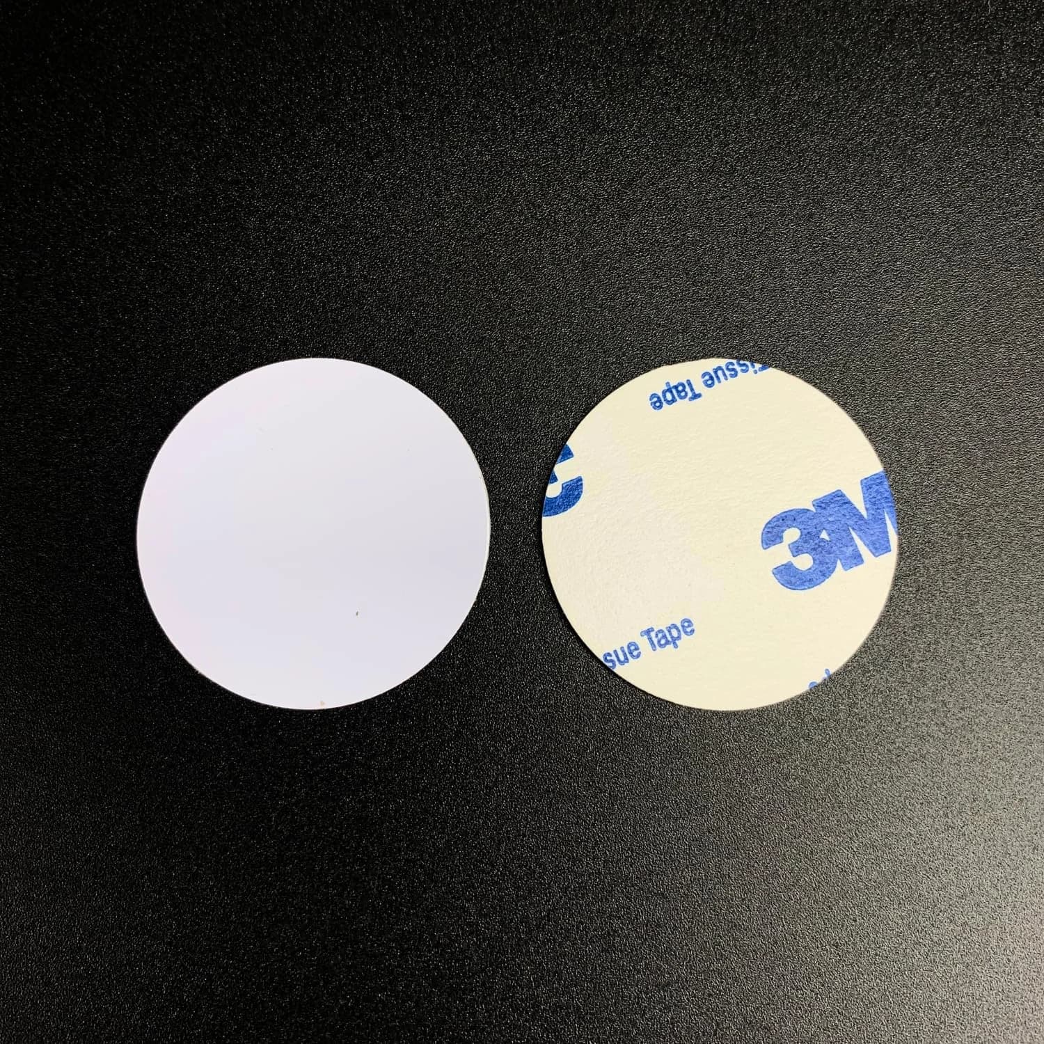 Two round white stickers on a black surface, one showing a 3M adhesive backing.