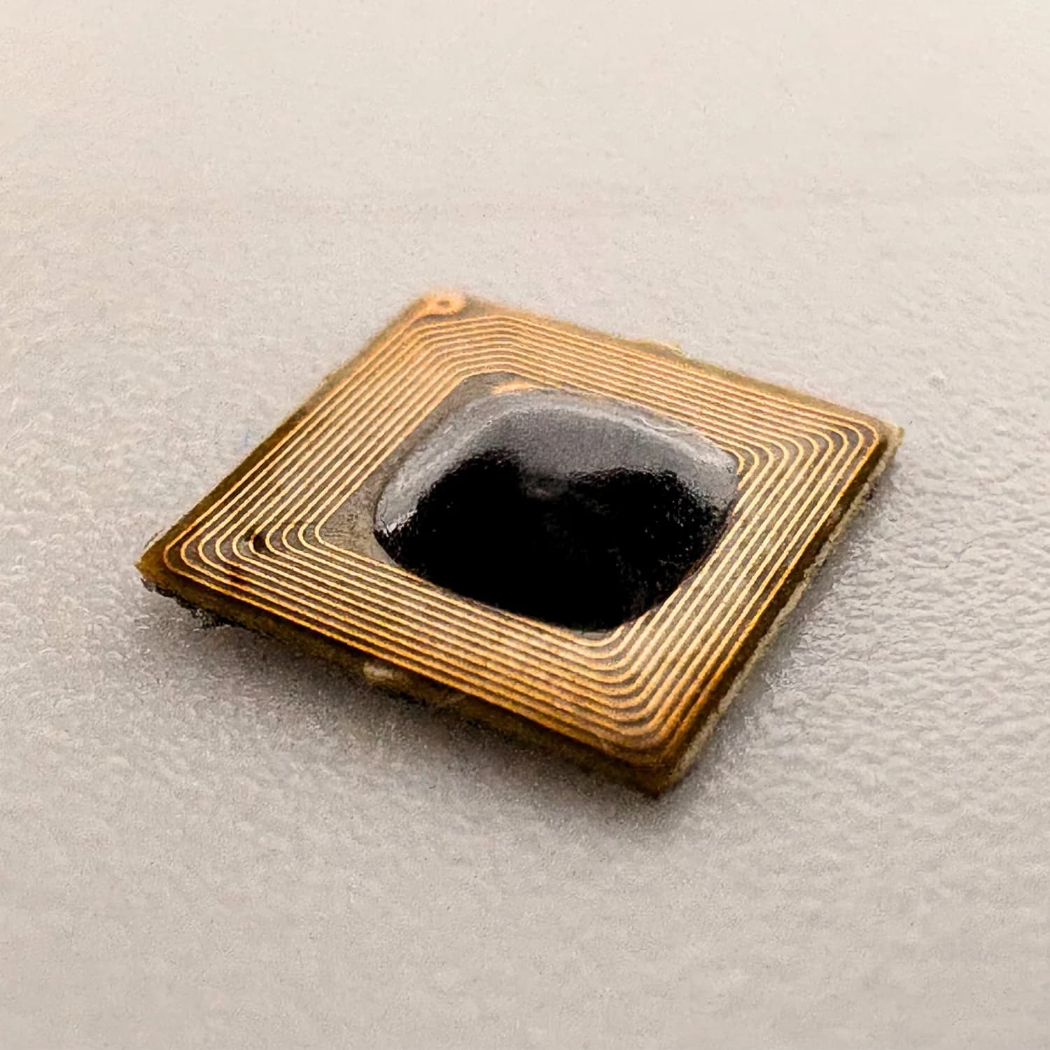 Close-up of a small, square NFC chip with gold-colored circuit lines on a gray background.